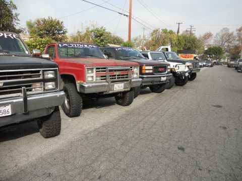 1986 Ford Bronco - A row of modified pickup trucks are parked on a street, with a churro stand visible in the background.