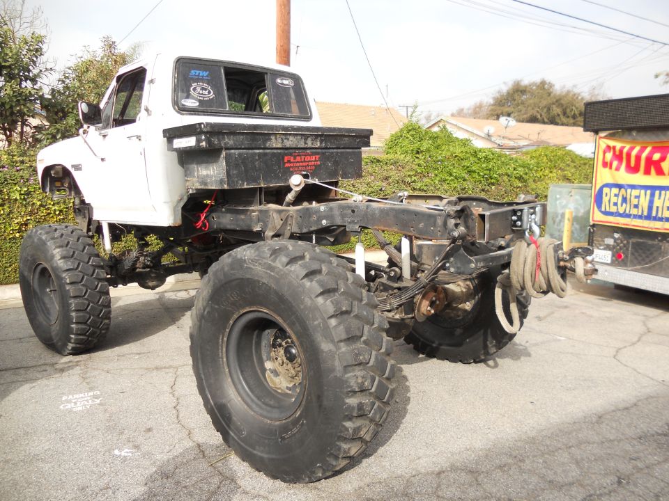 White pickup truck chassis with large off-road tires, a toolbox mounted on the frame, and a winch