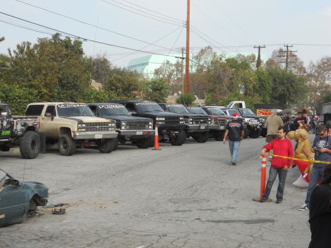 1986 Ford Bronco - A line of modified trucks and SUVs are parked in a lot, with people gathered around.
