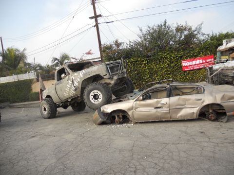 1986 Ford Bronco - A lifted monster truck is shown crushing a gold sedan, with a sign for 'Hobbies & Toys' visible in the background.