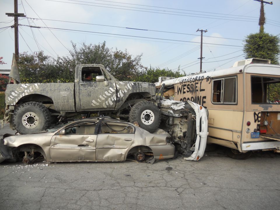 Mud-splattered pickup truck crushing a sedan, next to a bus with text