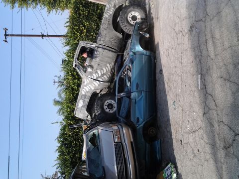 1986 Ford Bronco - A large gray pickup truck has crushed a teal sedan, with a third vehicle partially visible in the foreground.
