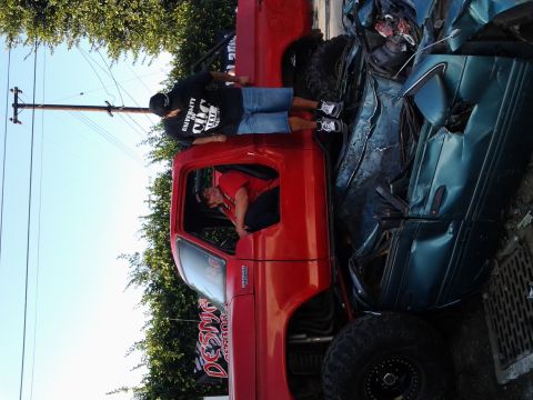 1986 Ford Bronco - A red pickup truck is parked beside a severely crushed teal car, with two people standing near the truck.
