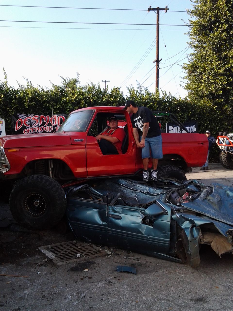 Red Ford pickup truck with large tires crushing a green sedan, two men standing by the truck