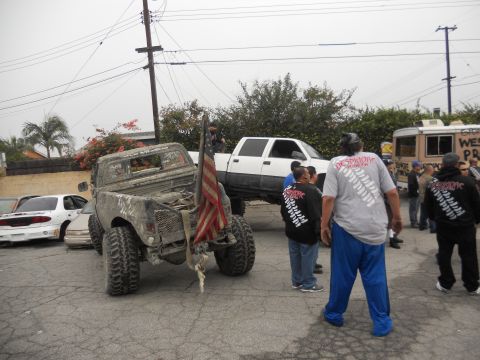 1986 Ford Bronco - A group of people gather around modified trucks, one with an American flag, on an overcast day.