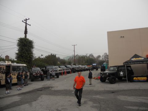 1986 Ford Bronco - A collection of modified trucks and SUVs are parked in a lot, with a man walking towards the camera.