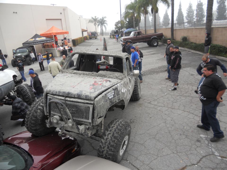 Mud-covered off-road truck with large tires, parked over a red car. Other vehicles and people are visible in the background