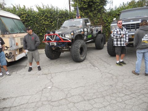 1986 Ford Bronco - A heavily modified black pickup truck with oversized tires and a roll cage is parked outdoors with other vehicles.