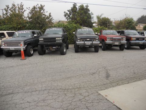 1986 Ford Bronco - A lineup of modified trucks and SUVs are parked on a cracked asphalt lot under an overcast sky.