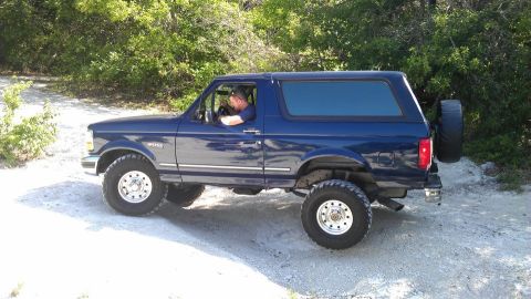 1995 Ford Bronco - A blue Ford Bronco is seen from the side on a dirt path surrounded by trees.