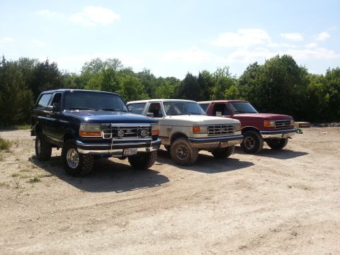 1995 Ford Bronco - Three SUVs, including a blue Ford Bronco, are parked side-by-side on a dusty lot with trees in the background.