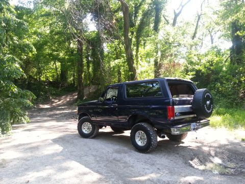 1995 Ford Bronco - A dark blue Ford Bronco is parked on a dirt path surrounded by trees.
