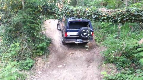 1995 Ford Bronco - 1995 Ford Bronco navigates a challenging off-road hill climb on a wooded trail.