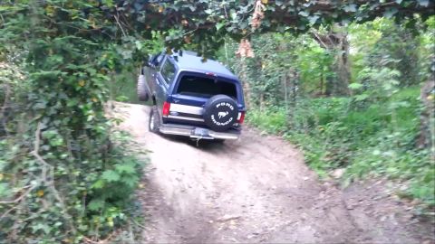 1995 Ford Bronco - A blue Ford Bronco navigates a steep, wooded trail, with its rear tire lifted off the ground.