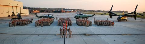 2000 Ford Crown Victoria - A formation of military personnel stands on an airfield with several helicopters and a hangar in the background.