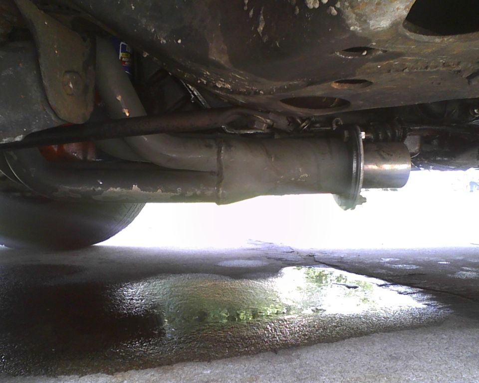 Underside view of a car's exhaust system, showing pipes and muffler. Puddles are visible on the ground below