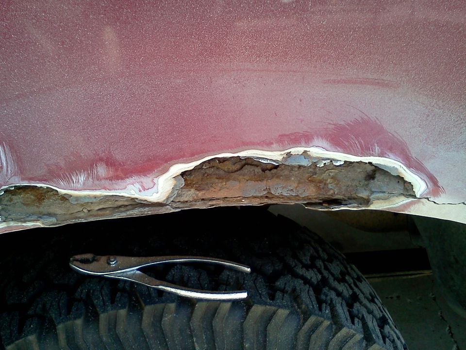 Rusted-through maroon fender arch above tire with pliers resting on tread