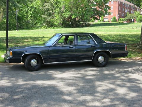 1991 Ford LTD Crown Victoria - dark navy-blue four-door sedan sits in profile on pavement next to a green lawn with brick apartment buildings behind.