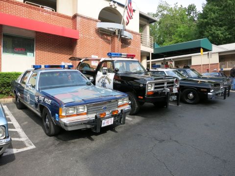 1991 Ford LTD Crown Victoria - blue Ford State Police sedan, a black Dodge truck and other vintage cruisers line up in a parking lot by a brick restaurant.