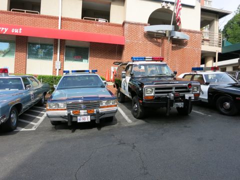 1991 Ford LTD Crown Victoria - blue Ford police sedan parks beside a black Dodge police pickup with winch and light bar in a lot by a brick building.