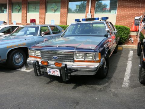 1991 Ford LTD Crown Victoria - blue Ford police sedan with chrome push bar and light bar parks nose-out beside another cruiser by a brick building.