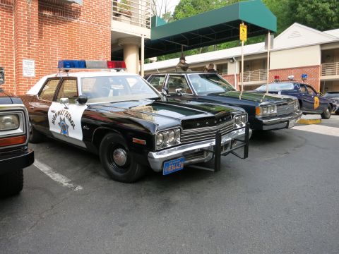 1991 Ford LTD Crown Victoria - black-and-white Dodge Highway Patrol sedan with push bar parks among other vintage cruisers in a lot by a brick motel.