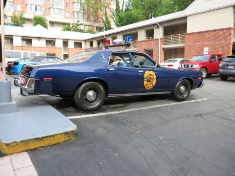 1991 Ford LTD Crown Victoria - dark blue classic Plymouth police sedan with a gold star door emblem and dome light parks in a lot among other cars.