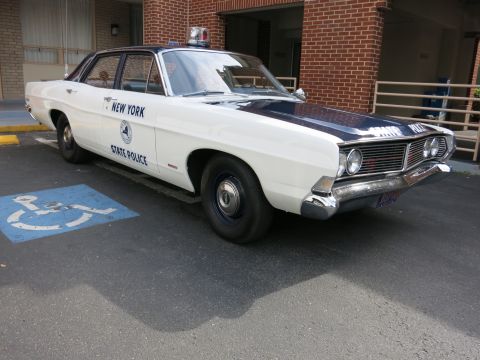 1991 Ford LTD Crown Victoria - white-and-black classic Ford New York State Police sedan with dome light sits in a handicap space beside a brick wall.