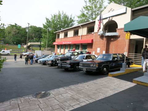 1991 Ford LTD Crown Victoria - long row of vintage police cars lines the front of a brick restaurant flying a US flag, with onlookers gathered nearby.