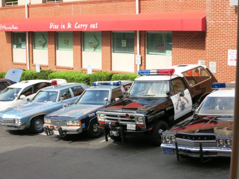 1991 Ford LTD Crown Victoria - Vintage police cars including a black Dodge truck and cruisers park nose-in against a brick restaurant with a red awning.