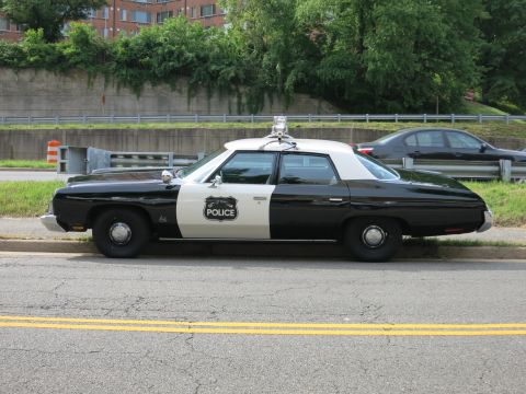 1991 Ford LTD Crown Victoria - vintage black-and-white police sedan with a roof beacon and POLICE door shield is parked on the shoulder near a retaining wall.