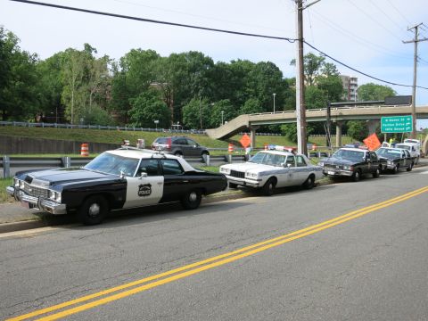 1991 Ford LTD Crown Victoria - Several vintage black-and-white and light-colored POLICE cruisers are lined up along a curb beside a highway overpass.
