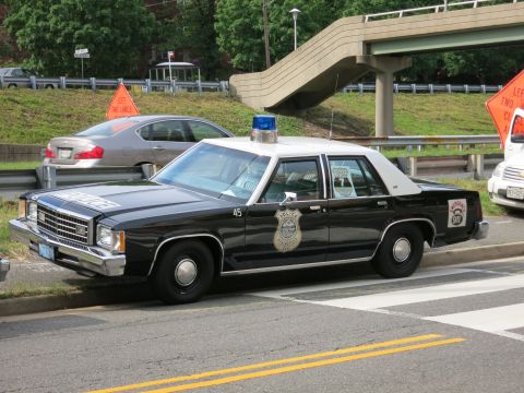 1991 Ford LTD Crown Victoria - black-and-white police sedan with a roof beacon, gold star door emblem, and number 45 is parked near a pedestrian overpass.