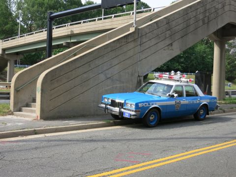 1991 Ford LTD Crown Victoria - blue-and-white police sedan marked POLICE with a roof lightbar sits beneath a concrete pedestrian overpass staircase.