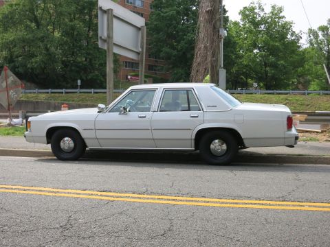 1991 Ford LTD Crown Victoria - plain white full-size sedan, side profile, parked at the curb on a tree-lined street near a building.