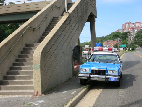 1991 Ford LTD Crown Victoria - front end of a blue police sedan with a roof lightbar and push bar is parked beside a concrete pedestrian overpass.