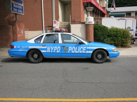 1991 Ford LTD Crown Victoria - blue full-size sedan lettered NYPD POLICE 103 PCT with a roof beacon is parked along the curb by a brick building.