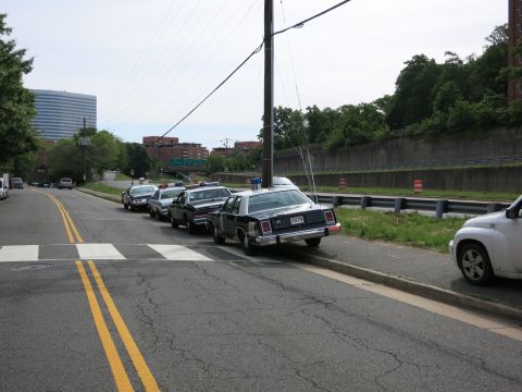 1991 Ford LTD Crown Victoria - line of dark vintage sedans is parked along a roadside curb with city buildings and a hillside in the background.