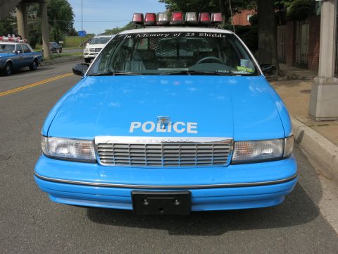 1991 Ford LTD Crown Victoria - Front view of a blue Chevrolet Caprice police car marked POLICE with a full roof lightbar and In Memory of 23 Shields windshield banner.