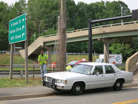 1991 Ford LTD Crown Victoria - white full-size sedan with a front push bar is parked near green highway signs while a worker in a hi-vis vest stands nearby.