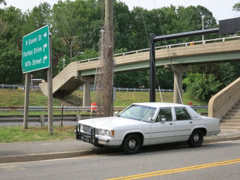 1991 Ford LTD Crown Victoria - white full-size sedan fitted with a front push bar is parked along a road by green highway signs and a concrete pedestrian overpass.