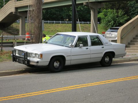 1991 Ford LTD Crown Victoria - white Ford full-size sedan with a chrome front push bar is parked at the roadside beneath a concrete pedestrian overpass.