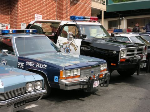 1991 Ford LTD Crown Victoria - cluster of police vehicles, including a blue STATE POLICE sedan, a highway-patrol car, and a black SUV, parked together near a brick buildi…