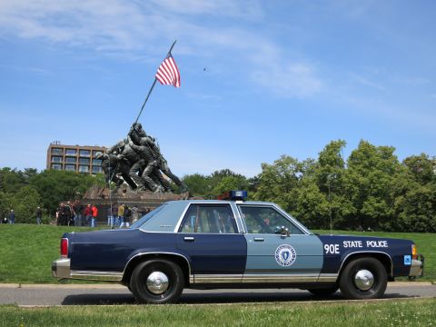1991 Ford LTD Crown Victoria - two-tone blue sedan marked 90E STATE POLICE is parked on grass in front of the Iwo Jima flag-raising memorial under a blue sky.