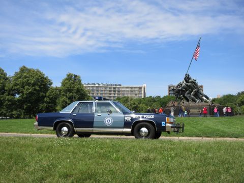 1991 Ford LTD Crown Victoria - close rear-quarter view of a two-tone blue 90E STATE POLICE sedan with the Iwo Jima flag-raising statue rising behind it.