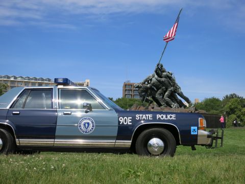 1991 Ford LTD Crown Victoria - vintage blue-and-silver Massachusetts State Police sedan marked 90E parked on grass in front of a bronze flag-raising war memorial.