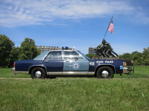1991 Ford LTD Crown Victoria - Driver-side profile of a blue-and-silver Massachusetts State Police sedan marked 90E parked on grass beside a war memorial under a clear sk…