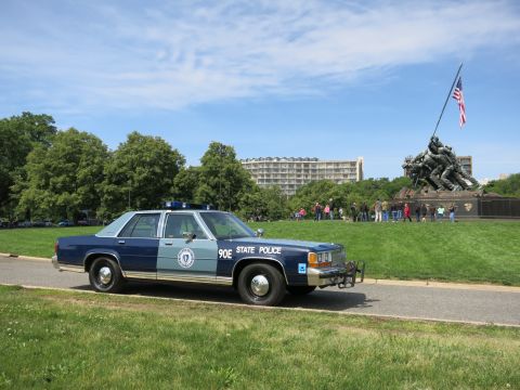 1991 Ford LTD Crown Victoria - blue-and-silver State Police sedan marked 90E parked on a path with a crowd gathered around a war memorial in the background.