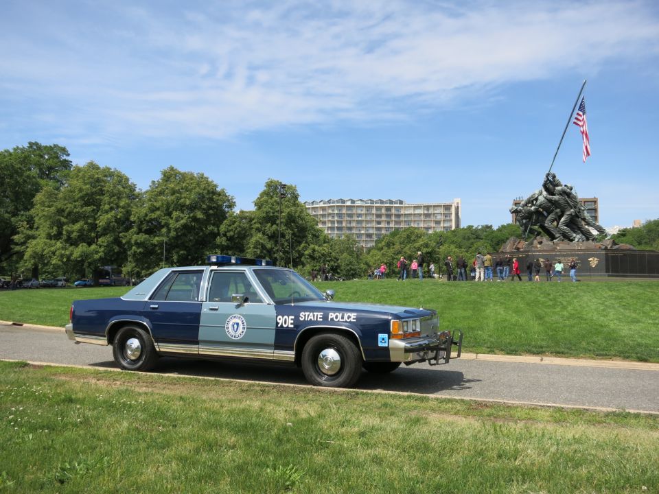 Blue and silver State Police sedan parked on a path with a crowd near a memorial behind