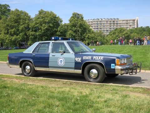 1991 Ford LTD Crown Victoria - Front-quarter view of a blue-and-silver State Police sedan marked 90E with a front push bar, parked on grass with onlookers behind.